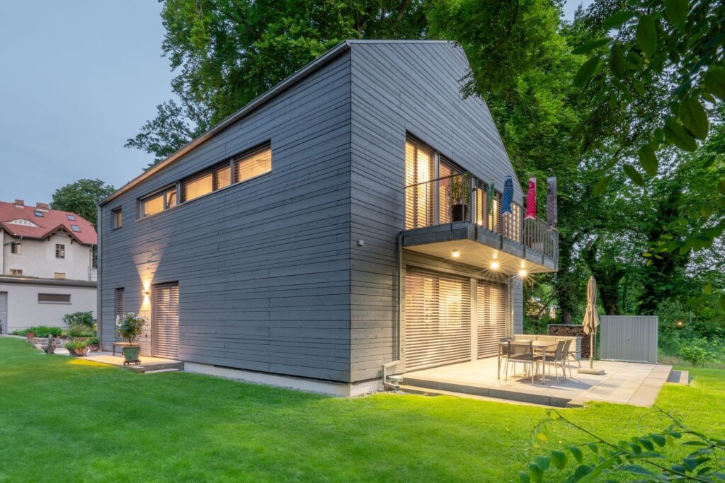 Haus mit grauer Gendai-Verkleidung vor Bäumen und mit Rasen